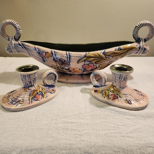 May include: A vintage ceramic centerpiece and candle holder set. The pieces are pink with blue and white accents, floral designs, and ring handles. The centerpiece is a boat shape, and the candle holders are shoe-shaped.