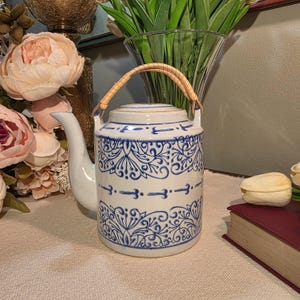 May include: A white ceramic teapot with blue floral patterns and a woven handle. The teapot has a long spout and a lid. The design includes a series of arrows and floral motifs. The teapot is a decorative item.