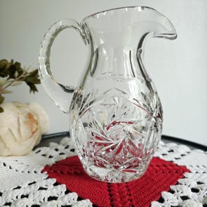 May include: A clear, cut-glass pitcher with a handle and a spout. The pitcher features a starburst pattern on the bottom half and is sitting on a red and white crocheted doily. The pitcher is likely used for serving beverages.