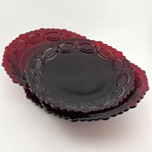 May include: Two stacked round plates made of deep red translucent glass. Each plate features a scalloped edge and a decorative border pattern consisting of embossed oval shapes and cross-hatched textures surrounding a smooth, circular center.