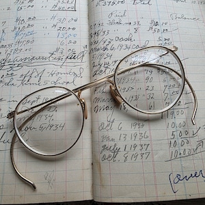 May include: A pair of vintage gold-rimmed eyeglasses rests on an open ledger filled with handwritten financial records. The glasses have round lenses and curved temples. The ledger's pages are aged and show faded ink.