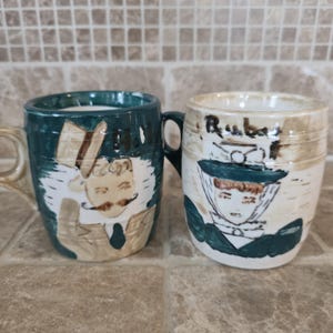 May include: A white ceramic mug with a black handle and the text "But First, Coffee" in a bold, black font. The mug is set against a blurred background, suggesting a cozy setting.