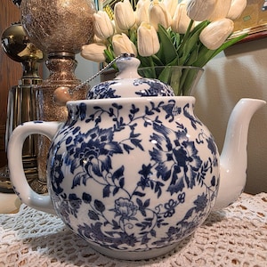 May include: White ceramic teapot with a blue floral pattern. The teapot has a curved spout, a rounded body, and a matching lid. The handle is a curved loop. The teapot is sitting on a white lace doily.