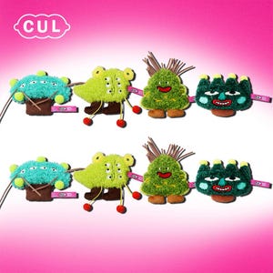 May include: A set of six plush toys with different designs. The toys are green, blue, and brown. Each toy has a different facial expression and features. The toys are all attached to a pink ribbon with the word "CUL" printed on it.