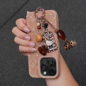 May include: A phone case with a floral pattern and a charm. The charm includes beads, a heart, and a Hello Kitty tag. The case is tan with a floral design and a camera cutout.