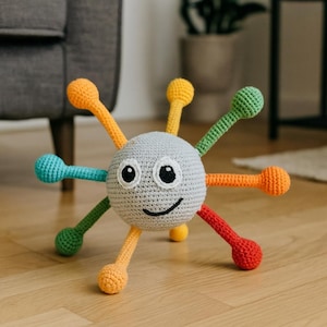 May include: A crocheted baby toy in the shape of a smiling face with multiple colorful arms. The toy has a gray head with black eyes and a curved mouth. The arms are in various colors, including orange, yellow, green, blue, and red.