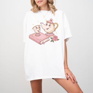 May include: White t-shirt with a cartoon illustration of a teapot and a teacup with a book and a rose. The teapot and teacup are from the Disney movie Beauty and the Beast.