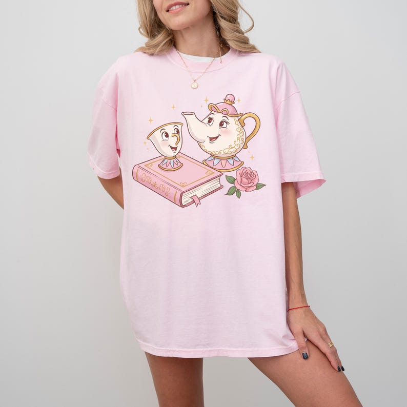 May include: A pink t-shirt with a cartoon image of a teapot and a teacup, both with faces. The teapot is pouring tea into the teacup. The image also includes a book with the word "Beauty" on the cover and a pink rose.