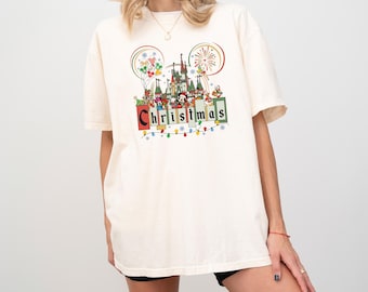 May include: Cream-colored t-shirt featuring a colorful Christmas design. The graphic includes a castle, Mickey Mouse ears, fireworks, and the word "Christmas" in a block letter style. The shirt is a relaxed fit.