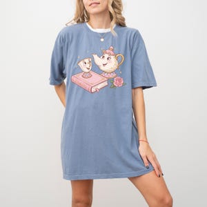 May include: A blue t-shirt with a graphic of a teapot and a teacup from the Disney movie Beauty and the Beast. The teapot is pink and white with a gold trim. The teacup is white with a gold trim. The graphic also includes a book and a rose.