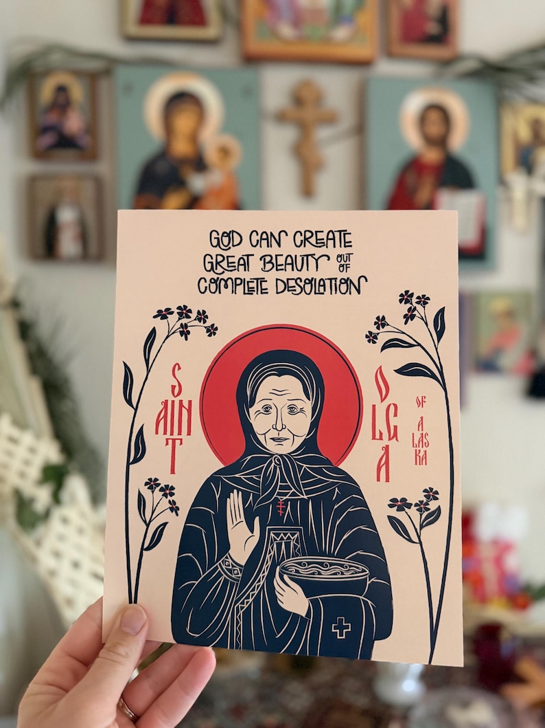Puede incluir: Una impresi&oacute;n rectangular sobre papel color melocot&oacute;n claro con una ilustraci&oacute;n de l&iacute;neas azul oscuro de Santa Olga de Alaska. El texto dice: GOD CAN CREATE GREAT BEAUTY OUT OF COMPLETE DESOLATION. La santa aparece con un halo rojo y tallos florales.