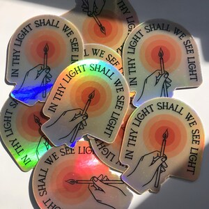 Puede incluir: Un conjunto de pegatinas hologr&aacute;ficas. Cada pegatina presenta una mano sosteniendo una vela, con un dise&ntilde;o de sol radiante en el fondo. El texto "IN THY LIGHT SHALL WE SEE LIGHT" est&aacute; impreso alrededor del borde.