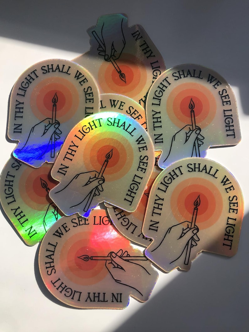 Puede incluir: Un conjunto de pegatinas hologr&aacute;ficas. Cada pegatina presenta una mano sosteniendo una vela, con un dise&ntilde;o de sol radiante en el fondo. El texto "IN THY LIGHT SHALL WE SEE LIGHT" est&aacute; impreso alrededor del borde.