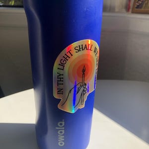 Puede incluir: Una botella de agua Owala azul con tapa turquesa. Una pegatina hologr&aacute;fica est&aacute; adherida a la botella, con una vela y el texto "IN THY LIGHT SHALL WE SEE LIGHT". La botella est&aacute; sobre una superficie blanca.