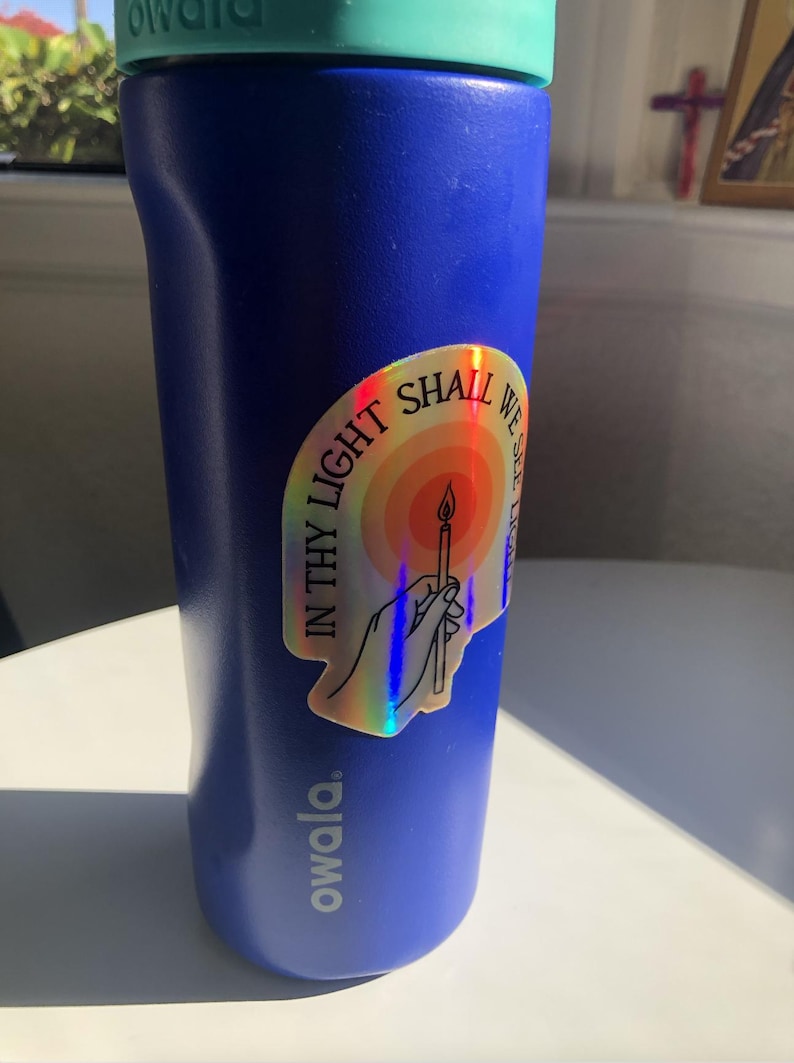 Puede incluir: Una botella de agua Owala azul con tapa turquesa. Una pegatina hologr&aacute;fica est&aacute; adherida a la botella, con una vela y el texto "IN THY LIGHT SHALL WE SEE LIGHT". La botella est&aacute; sobre una superficie blanca.