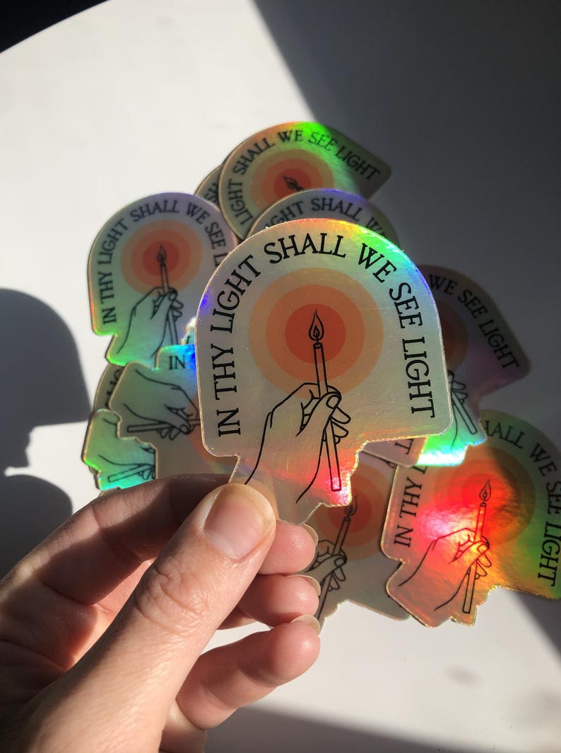 Puede incluir: Pegatinas hologr&aacute;ficas con un dise&ntilde;o que presenta una mano sosteniendo una vela, rodeada de un sol en tonos c&aacute;lidos. El texto "IN THY LIGHT SHALL WE SEE LIGHT" est&aacute; impreso en las pegatinas. Las pegatinas tienen un brillo arco&iacute;ris.