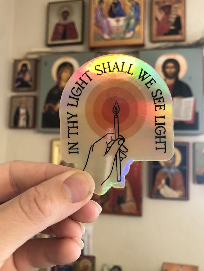 Puede incluir: Una pegatina hologr&aacute;fica con el texto "IN THY LIGHT SHALL WE SEE LIGHT" en un arco iris. La pegatina presenta una mano sosteniendo una vela encendida sobre un fondo de c&iacute;rculos conc&eacute;ntricos en tonos naranja y amarillo.