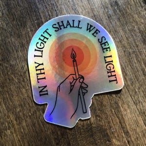 Puede incluir: Pegatina hologr&aacute;fica con el texto "IN THY LIGHT SHALL WE SEE LIGHT" en semic&iacute;rculo. La pegatina presenta una mano sosteniendo una vela encendida sobre un fondo de arco&iacute;ris.