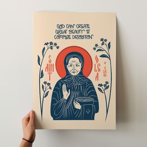 Puede incluir: Una impresi&oacute;n rectangular sobre fondo beige claro con una ilustraci&oacute;n azul oscuro de Santa Olga de Alaska. El texto dice: GOD CAN CREATE GREAT BEAUTY OUT OF COMPLETE DESOLATION. La figura tiene un halo rojo y est&aacute; flanqueada por flores azules.
