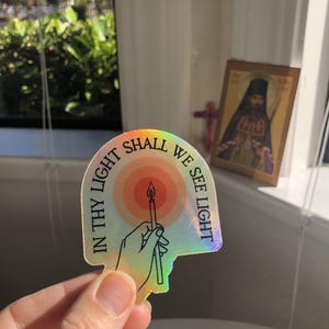 Puede incluir: Pegatina hologr&aacute;fica con el texto "IN THY LIGHT SHALL WE SEE LIGHT" alrededor de una mano que sostiene una vela encendida. La pegatina tiene un brillo de arco&iacute;ris y un dise&ntilde;o estilizado. Un icono religioso es visible en el fondo.