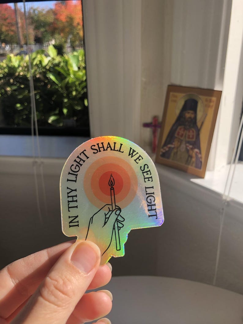 Puede incluir: Pegatina hologr&aacute;fica con el texto "IN THY LIGHT SHALL WE SEE LIGHT" alrededor de una mano que sostiene una vela encendida. La pegatina tiene un brillo de arco&iacute;ris y un dise&ntilde;o estilizado. Un icono religioso es visible en el fondo.