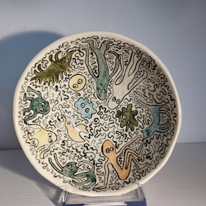 May include: A white ceramic bowl with a hand-painted, whimsical design. The bowl features various colourful, cartoonish creatures and shapes outlined in black against a white background. The bowl is displayed on a clear plastic stand.