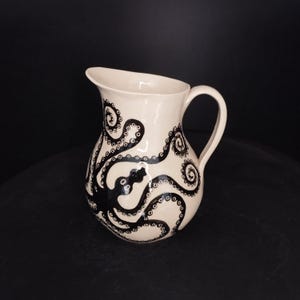 May include: Cream ceramic jug featuring a black octopus design. The tentacles of the octopus are wrapped around the jug's body, creating a striking contrast. The jug has a curved handle and a pouring spout.