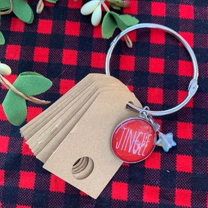 Peut inclure: Un ensemble d'étiquettes cadeaux brunes avec un trou perforé en haut, attaché à un anneau argenté avec un charme rond rouge qui dit "Jingle" et un charme étoile argenté.