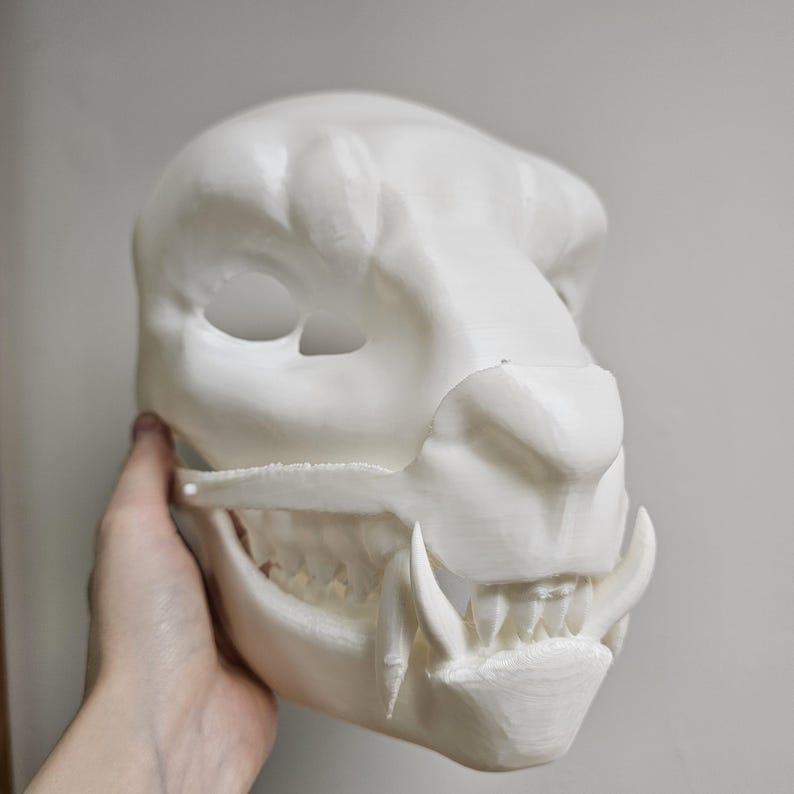 May include: A white, 3D-printed mask of a feline skull, featuring detailed teeth and a textured surface. The mask has eye openings and a prominent jawline, suggesting a realistic design. The mask is held by a hand.