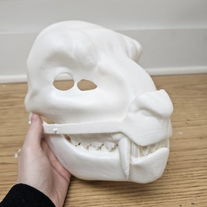 May include: A white, 3D-printed mask in the shape of a snarling beast's head. The mask features eye holes, a prominent snout, sharp teeth, and a detailed jawline. The mask is held by a hand.