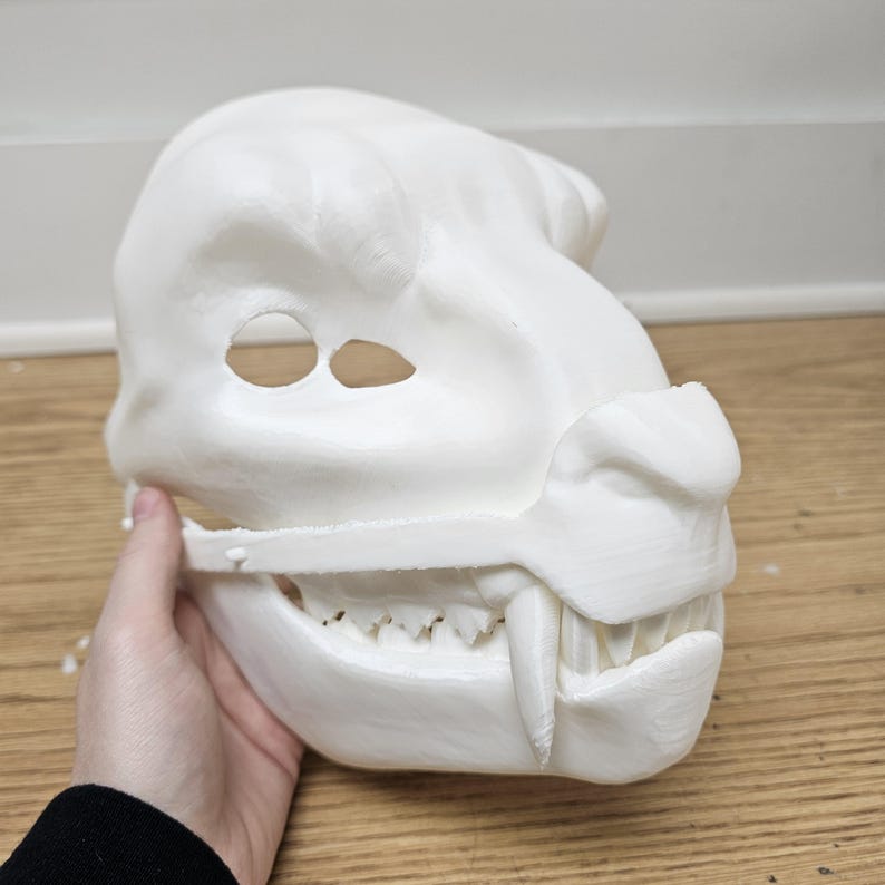 May include: A white, 3D-printed mask in the shape of a snarling beast's head. The mask features eye holes, a prominent snout, sharp teeth, and a detailed jawline. The mask is held by a hand.