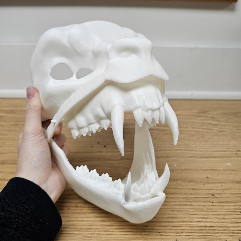 May include: A white, 3D-printed mask shaped like a ferocious animal skull. The mask features large, sharp teeth and a detailed bone structure, perfect for cosplay or Halloween. The mask is held by a person.