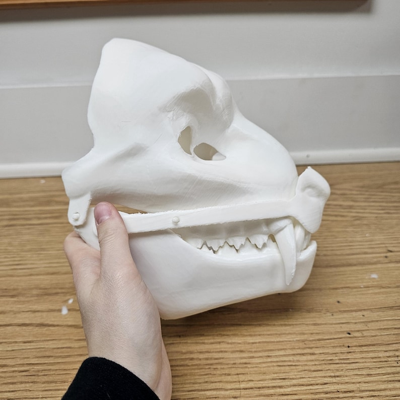 May include: A white, 3D-printed mask of a canine or bear-like creature. The mask features detailed teeth, a prominent snout, and a hinged jaw. The mask is held by a person's hand, showcasing its size and design.