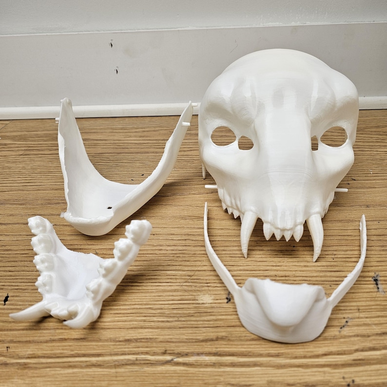 May include: A white 3D-printed cat skull mask with separate jaw and teeth components. The mask features large eye sockets and prominent fangs, suggesting a fantasy or Halloween theme. The pieces are arranged on a wooden surface.