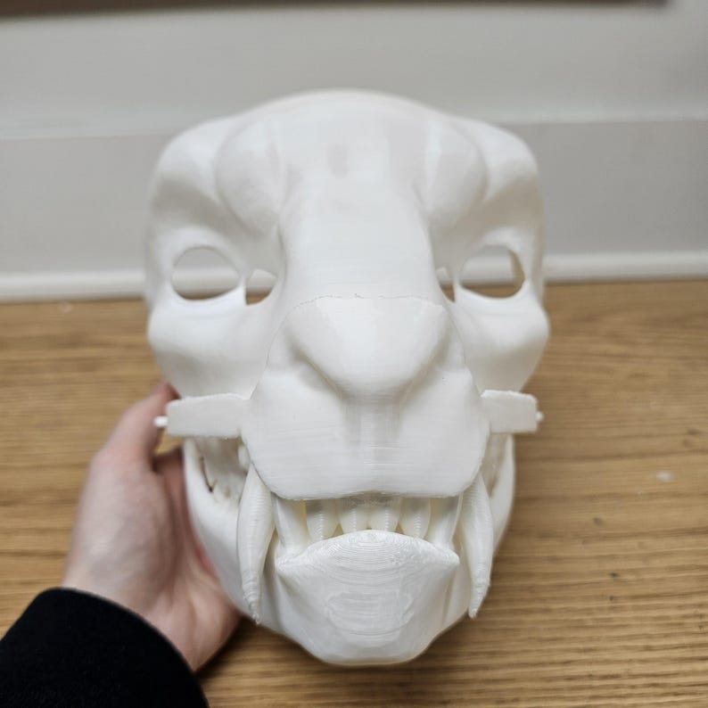 May include: A white, 3D-printed mask of a feline skull, featuring prominent canines and eye sockets. The mask is held by a hand, showcasing its detailed design and texture. The mask is likely for cosplay or costume use.