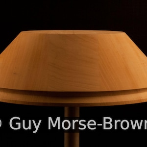 May include: A wooden lampshade with a light brown finish. The lampshade has a wide, circular shape and a slightly curved top.