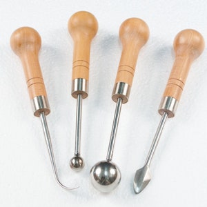 May include: Set of four wooden handled sculpting tools with metal tips. The tools have different shapes, including a round ball, a pointed tip, and a curved tip.