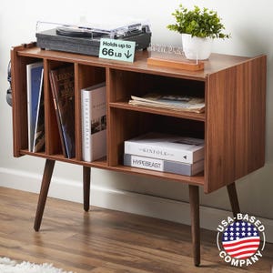 May include: A wooden record player stand with a turntable on top, shelves for records and books, and a sign that says "Now Playing". The stand has a mid-century modern design with tapered legs. The stand is made by a USA-based company.
