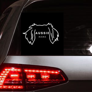 Peut inclure: Une voiture noire avec un autocollant blanc sur la vitre arrière. L'autocollant représente la silhouette d'un Berger Australien avec le texte "Aussie Mama" en dessous.