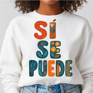 May include: White sweatshirt with the phrase "Sí Se Puede" in colorful, decorative lettering. The text features orange, teal, and floral designs. The sweatshirt is a casual, comfortable style.