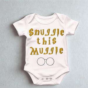 May include: White baby onesie with the text "Snuggle this Muggle" in gold lettering and a pair of black-rimmed glasses. The onesie is a short-sleeved garment with snap closures at the bottom, designed for infants.