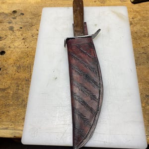 May include: A fixed-blade knife with a wooden handle and a metal crossguard, housed in a dark brown leather sheath. The sheath features a decorative embossed pattern of diagonal feather-like motifs and is stitched along the outer edges.