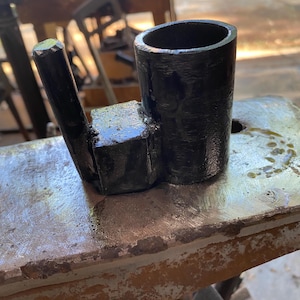 May include: A black metal tool with a cylindrical cup and a handle attached to a square base. The tool is sitting on a metal surface with a worn, textured appearance. The background is blurred, suggesting an outdoor workshop setting.