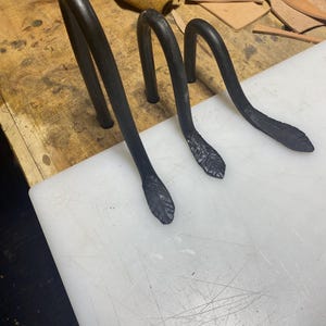 May include: Three black metal hooks with a curved design. The hooks have a leaf-like design at the base. The hooks are displayed on a white cutting board with a wooden surface in the background.