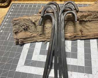 May include: A collection of hand-forged, dark metal hooks with curved tops and pointed ends. The hooks are arranged together on a piece of weathered wood, set against a gray cutting mat with measurement markings.