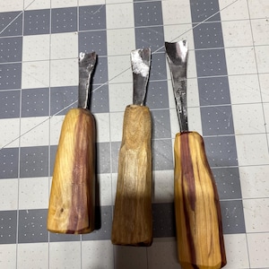 May include: Three wood carving gouges with metal blades. The handles are made of light-colored wood with reddish-brown streaks. The blades are silver and have a curved shape, designed for detailed woodworking.