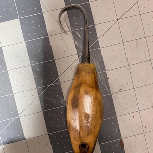May include: A wood-handled pottery trimming tool with a curved metal blade. The handle is a light brown color with a glossy finish. The blade is curved and appears to be made of metal. The tool is designed for shaping and refining clay.