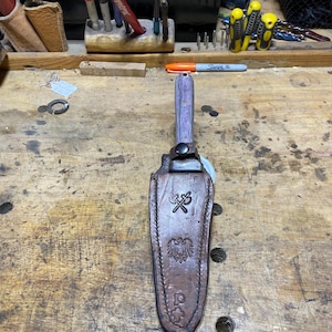 May include: A handcrafted dagger with a purple and gray handle and a brown leather sheath. The sheath features embossed designs including an eagle and a cross. The dagger is resting on a wooden workbench.
