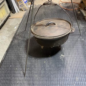 May include: A black cast iron Dutch oven with a lid and handle, suspended from a tripod. The pot is round with a curved handle on the lid. The tripod is made of metal and has a chain for hanging the pot over a fire.