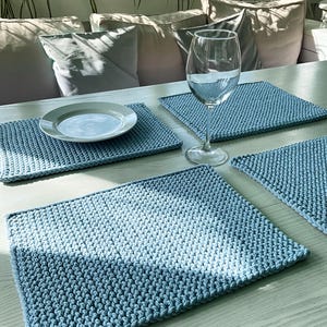 Könnte beinhalten: Vier hellblaue gehäkelte Tischsets auf einem hellen Holztisch. Ein weißer Teller und ein Weinglas stehen auf dem Tisch. Die Tischsets haben ein strukturiertes, gewebtes Aussehen. Der Hintergrund umfasst Kissen und Grün.